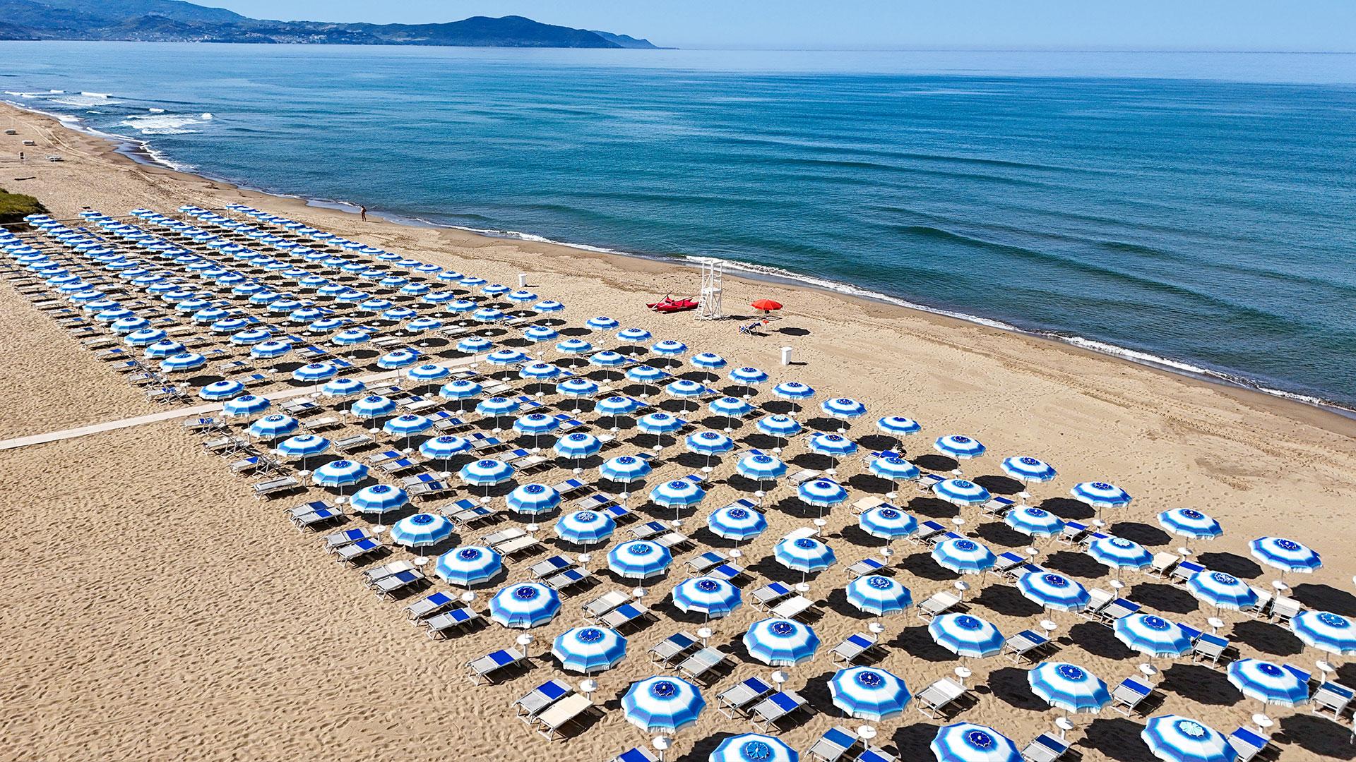 Spiaggia con file di ombrelloni blu e bianchi, mare calmo e montagne sullo sfondo.