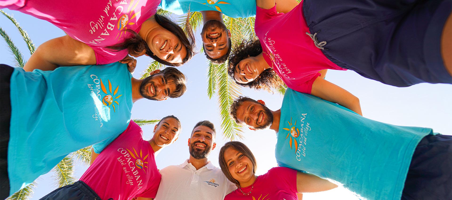 Gruppo di persone sorridenti in cerchio, indossano magliette colorate e palme sullo sfondo.