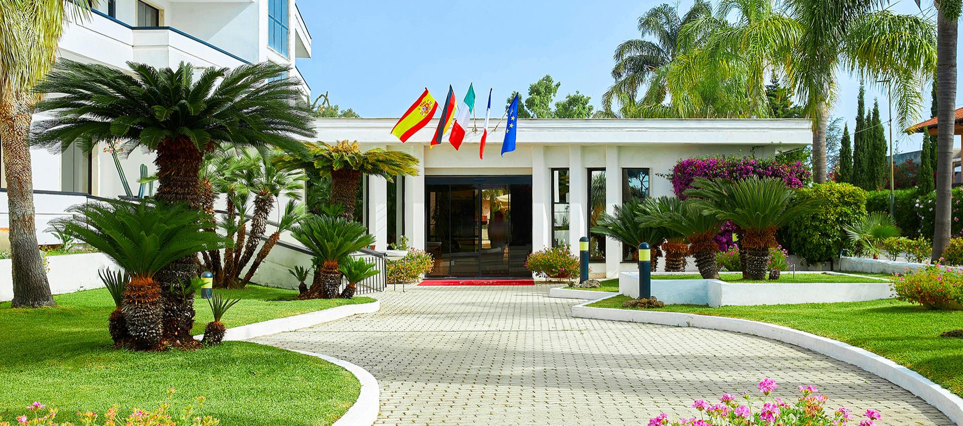 Ingresso elegante con bandiere e giardino curato, circondato da palme e fiori.