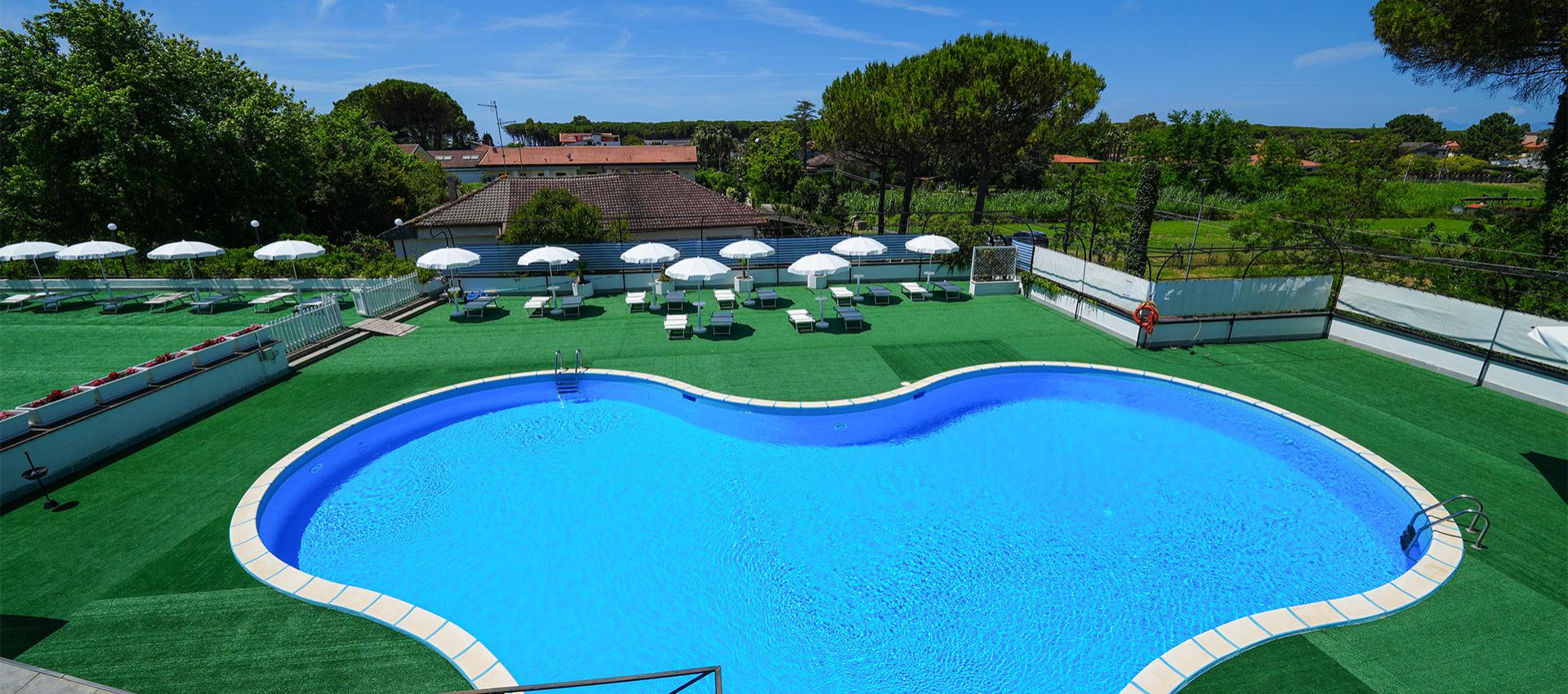 Piscina a forma libera, circondata da lettini e ombrelloni su prato sintetico verde.