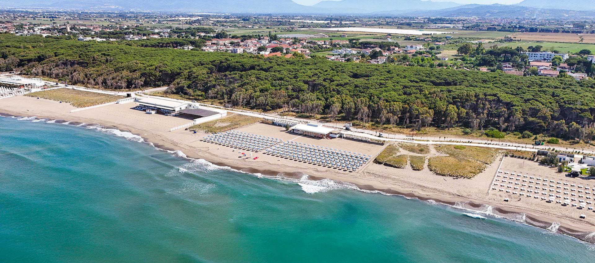 Spiaggia con ombrelloni e mare limpido, circondata da una fitta pineta e paesaggi rurali.