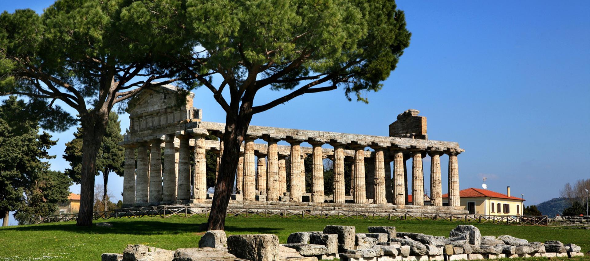 Tempio antico con colonne doriche, circondato da alberi e rovine, sotto un cielo azzurro.