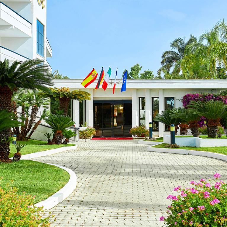 Ingresso di un edificio con bandiere e giardino ben curato, palme e fiori.