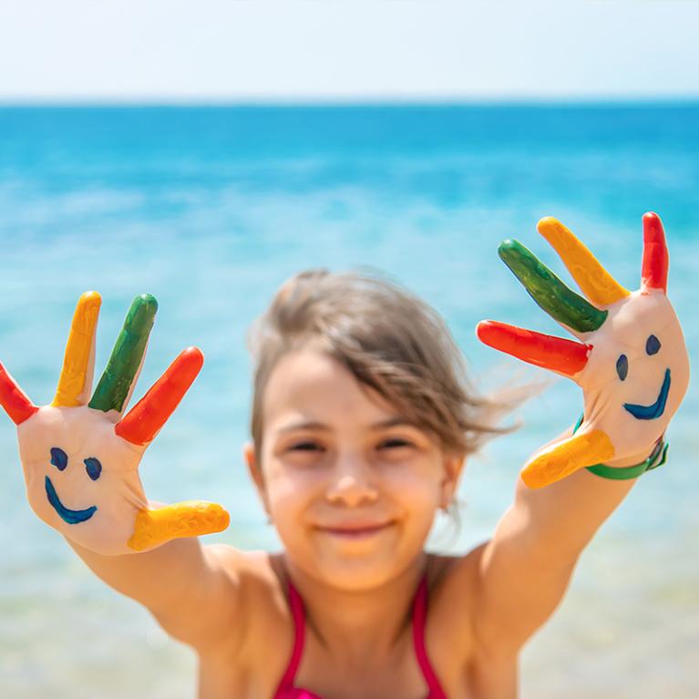 Bambina sorridente con mani dipinte colorate, sfondo mare.