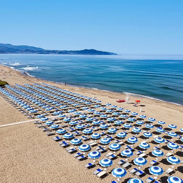 Spiaggia con ombrelloni blu e bianchi allineati, mare calmo e montagne sullo sfondo.