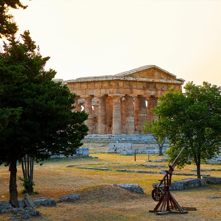 Tempio greco antico circondato da alberi, con una catapulta in primo piano.