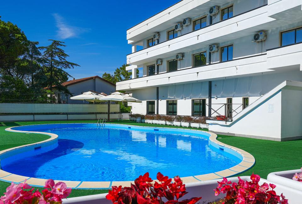 Piscina all'aperto con area verde, fiori e edificio bianco sullo sfondo.