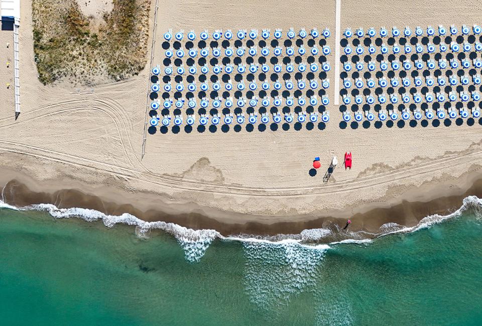 Spiaggia con file di ombrelloni blu e mare cristallino, vista dall'alto.