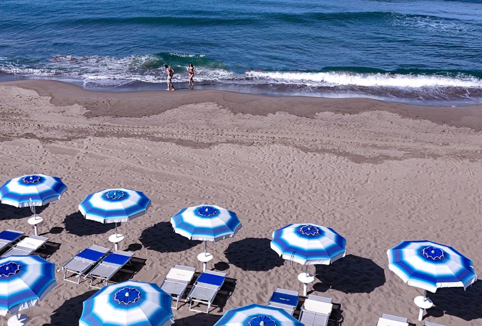 Spiaggia con ombrelloni blu e bianchi, due persone camminano lungo la riva.