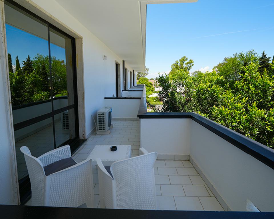 Balcone moderno con sedie bianche, vista su alberi verdi e cielo azzurro.